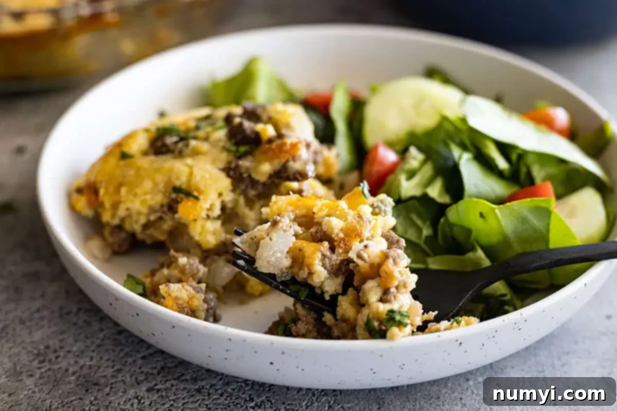 A serving of impossible cheeseburger pie in a white shallow bowl with a fork taking a bite out. 