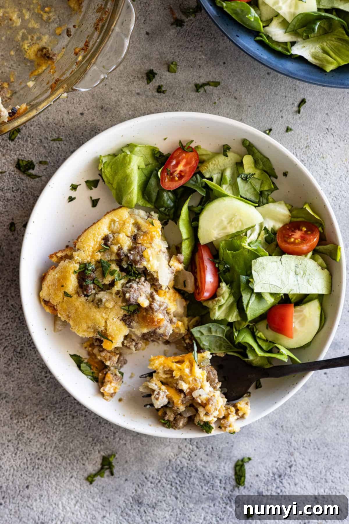 Overhead view of a slice of impossible cheeseburger pie in a shallow white bowl with a salad. Fork in the plate. 