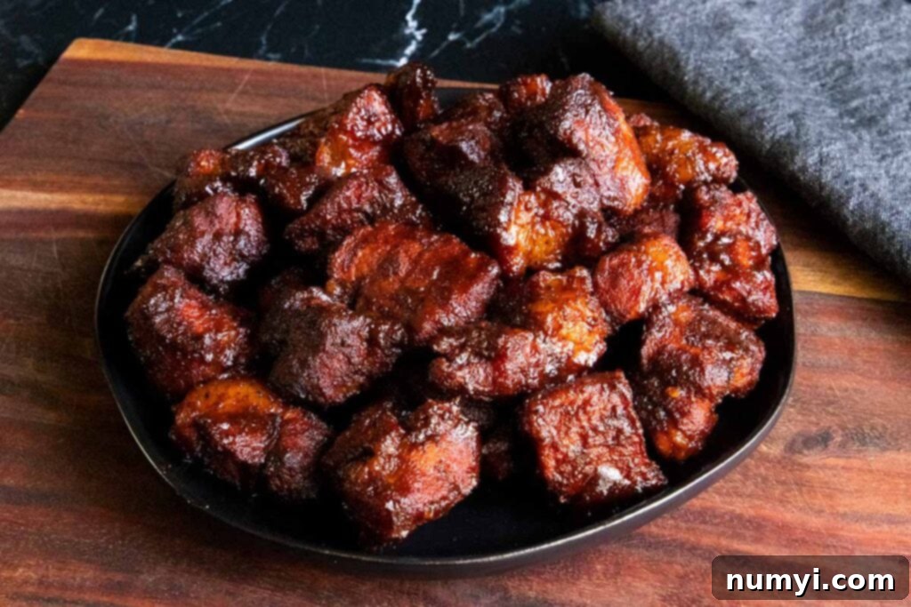 Ultimate Pork Butt Burnt Ends 2 Pork butt burnt ends piled high on a black plate, resting on a rustic wooden board, showcasing their glossy, dark bark and tender texture.