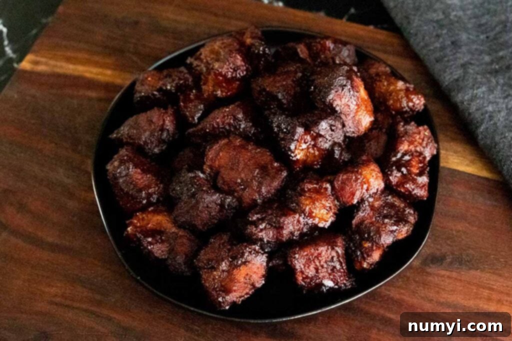 Ultimate Pork Butt Burnt Ends 17 A close-up of glossy, dark pork butt burnt ends artfully arranged on a black plate, emphasizing their texture and appealing glaze.