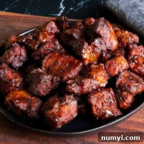 Ultimate Pork Butt Burnt Ends 18 Pork butt burnt ends piled on a black plate on top of wooden board.