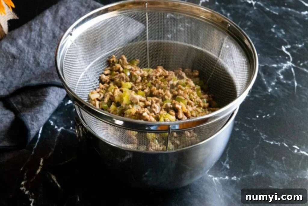 Meat mixture for dirty rice being drained in a strainer set over a bowl, showing excess liquid separating from the rich, savory meat, ensuring a non-greasy final dish.