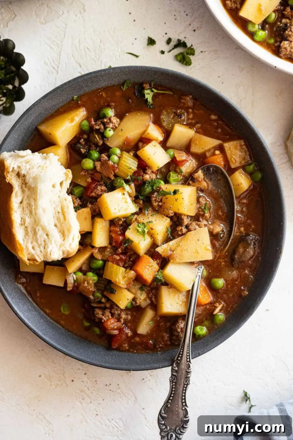 Overhead view of stew in a dark gray bowl with spoon in the soup and biscuit off to the side. 
