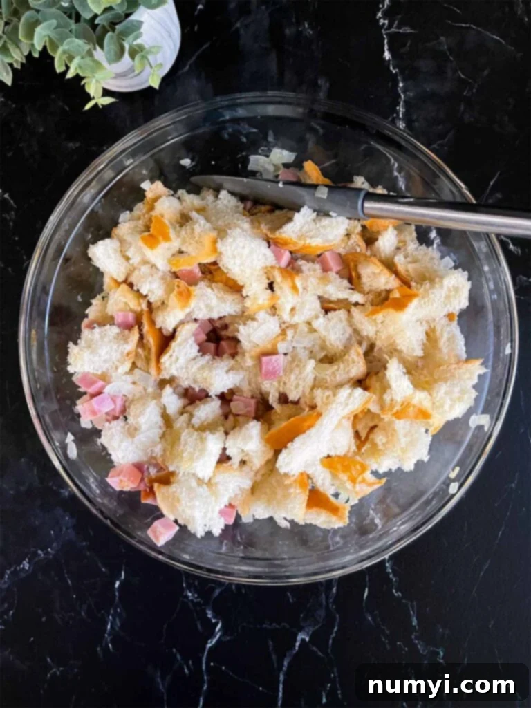 A mix of torn bread pieces, caramelized onions, and diced ham in a large glass mixing bowl on a dark surface, ready for the egg custard.