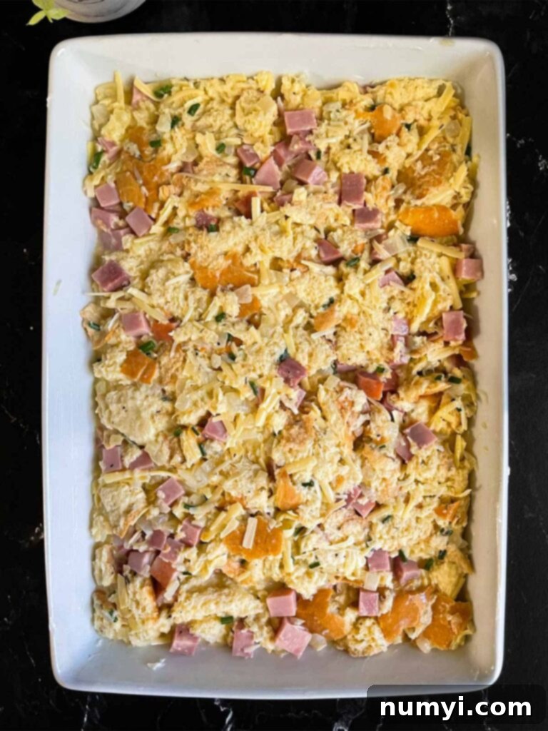 An unbaked ham and cheese strata, covered with plastic wrap, patiently waiting in a baking dish after an overnight soak.