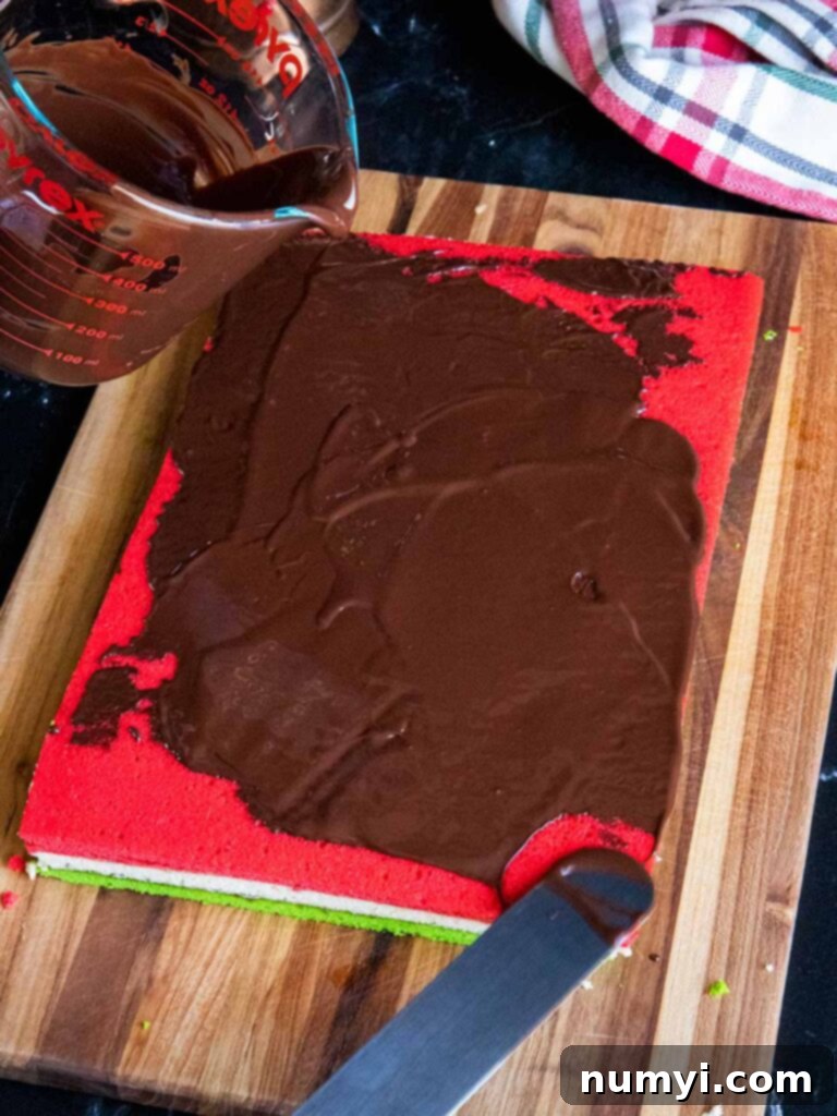 Smooth melted chocolate being evenly spread over the red top layer of the Italian rainbow cookie slab, creating a sleek and decadent coating.