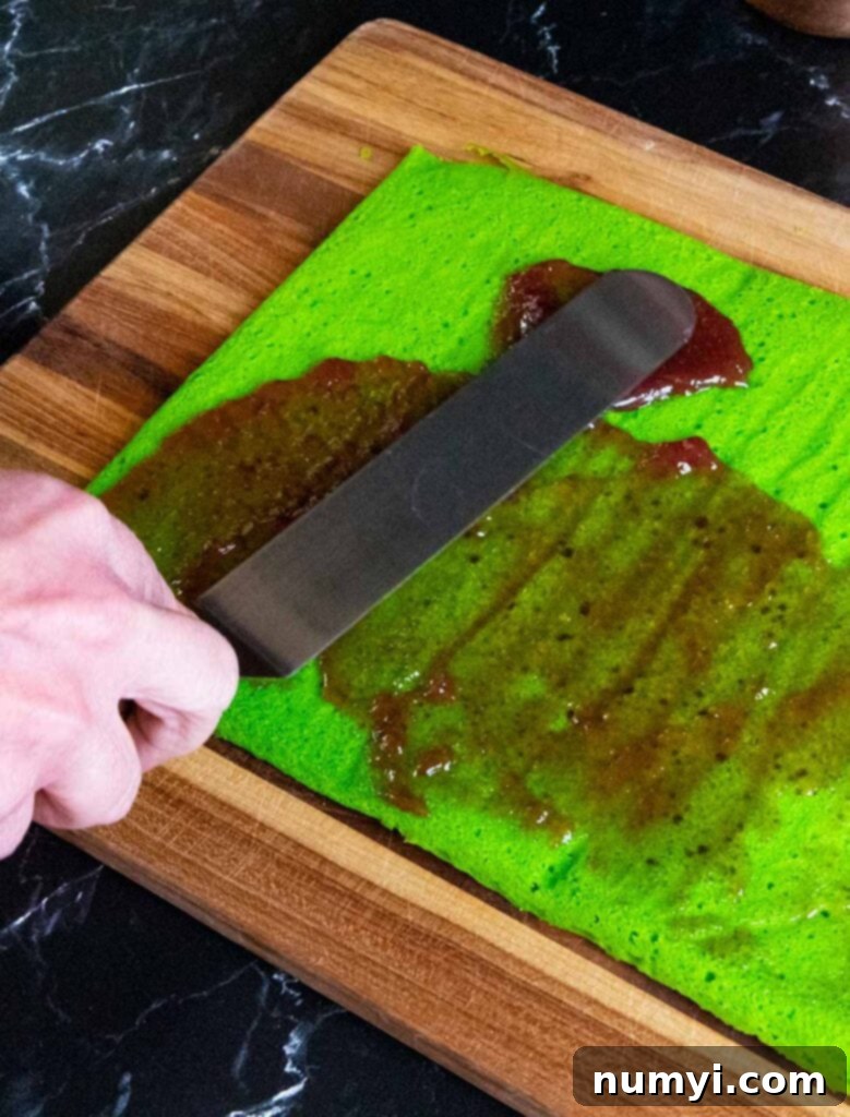 Sweet raspberry jam being meticulously spread over the vibrant green layer of an Italian rainbow cookie, creating a delicious and colorful filling.