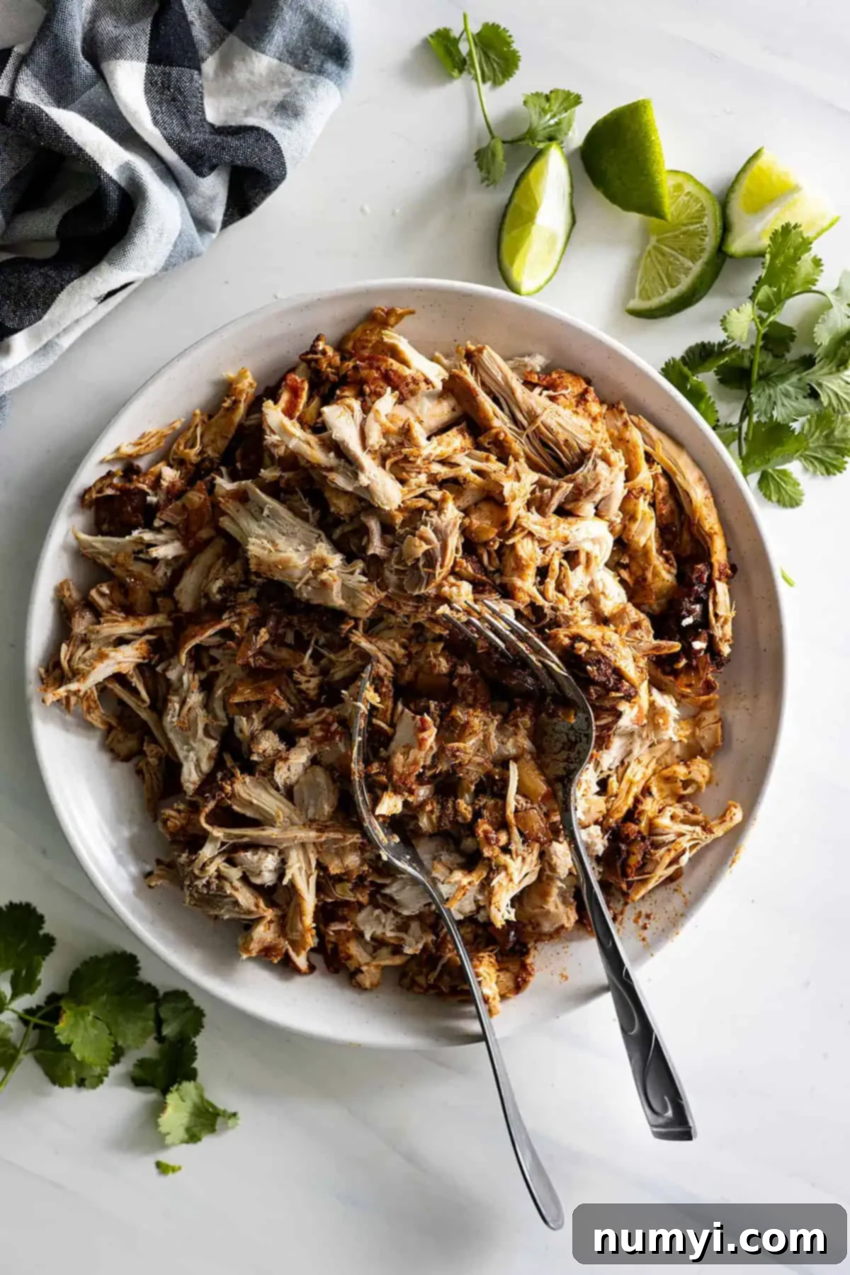 Overhead view of chicken taco filling in a white bowl with forks in it and cilantro and limes off to the side. 