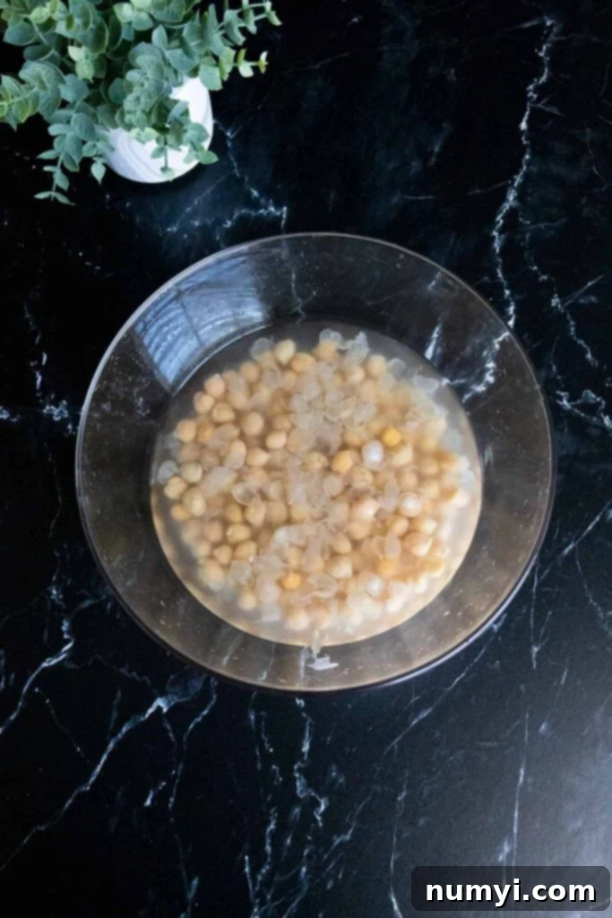 Rinsed chickpeas soaking in a glass bowl filled with hot water, preparing for skin removal.