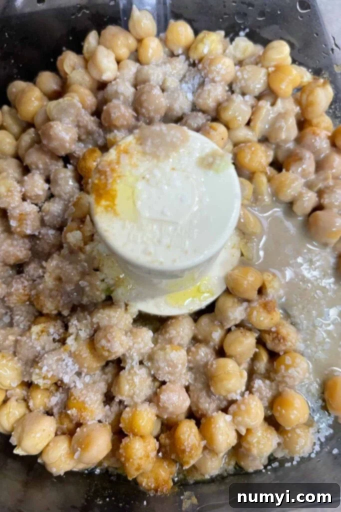 All hummus ingredients including chickpeas, garlic, lemon, tahini, cumin, and olive oil, in the bowl of a food processor.