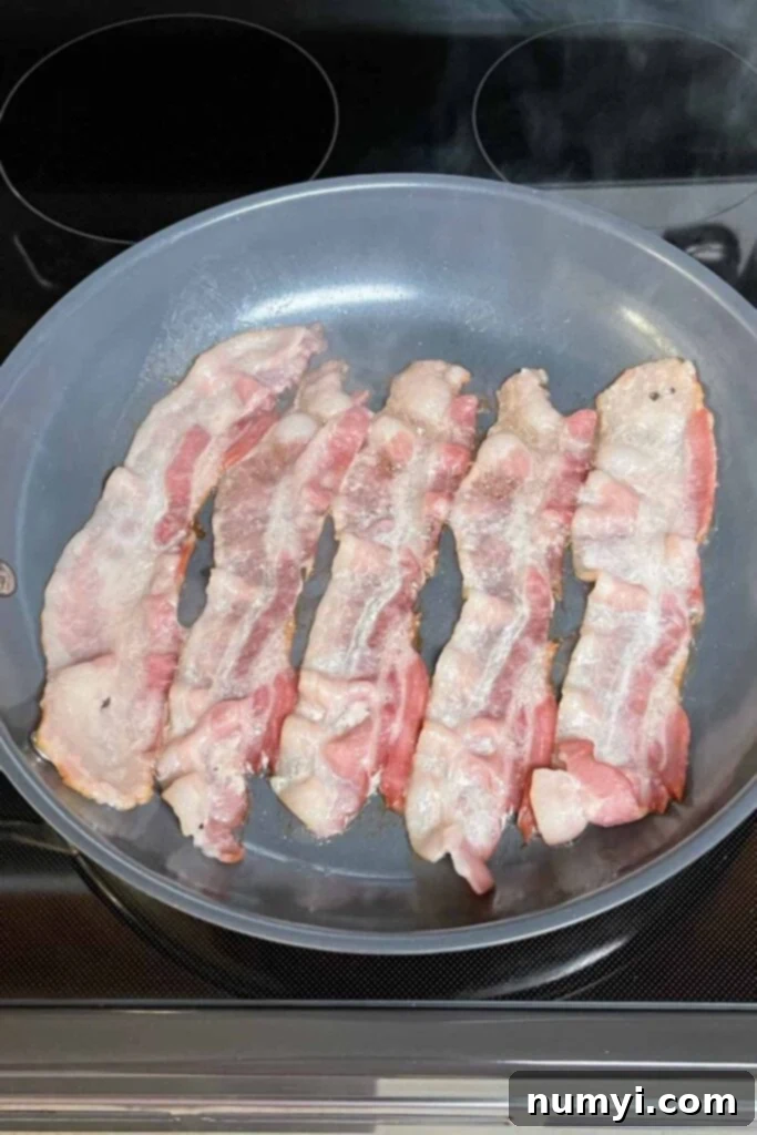 Bacon cooking in a pan until crispy, with reserved bacon grease.