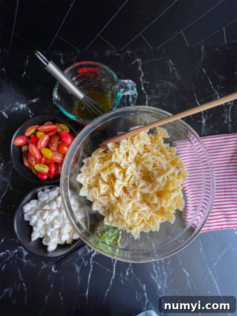 Cooked pasta in a glass mixing bowl, pearl mozzarella in a small black bowl, halved grape tomatoes in a small black bowl, and the vibrant pesto vinaigrette in a measuring cup, all ready for assembly.