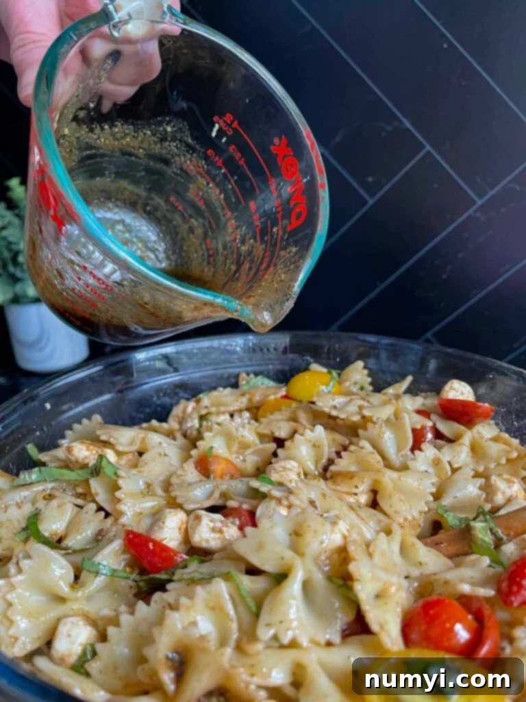 Vinaigrette dressing being drizzled over the colorful Caprese pasta salad in a mixing bowl.