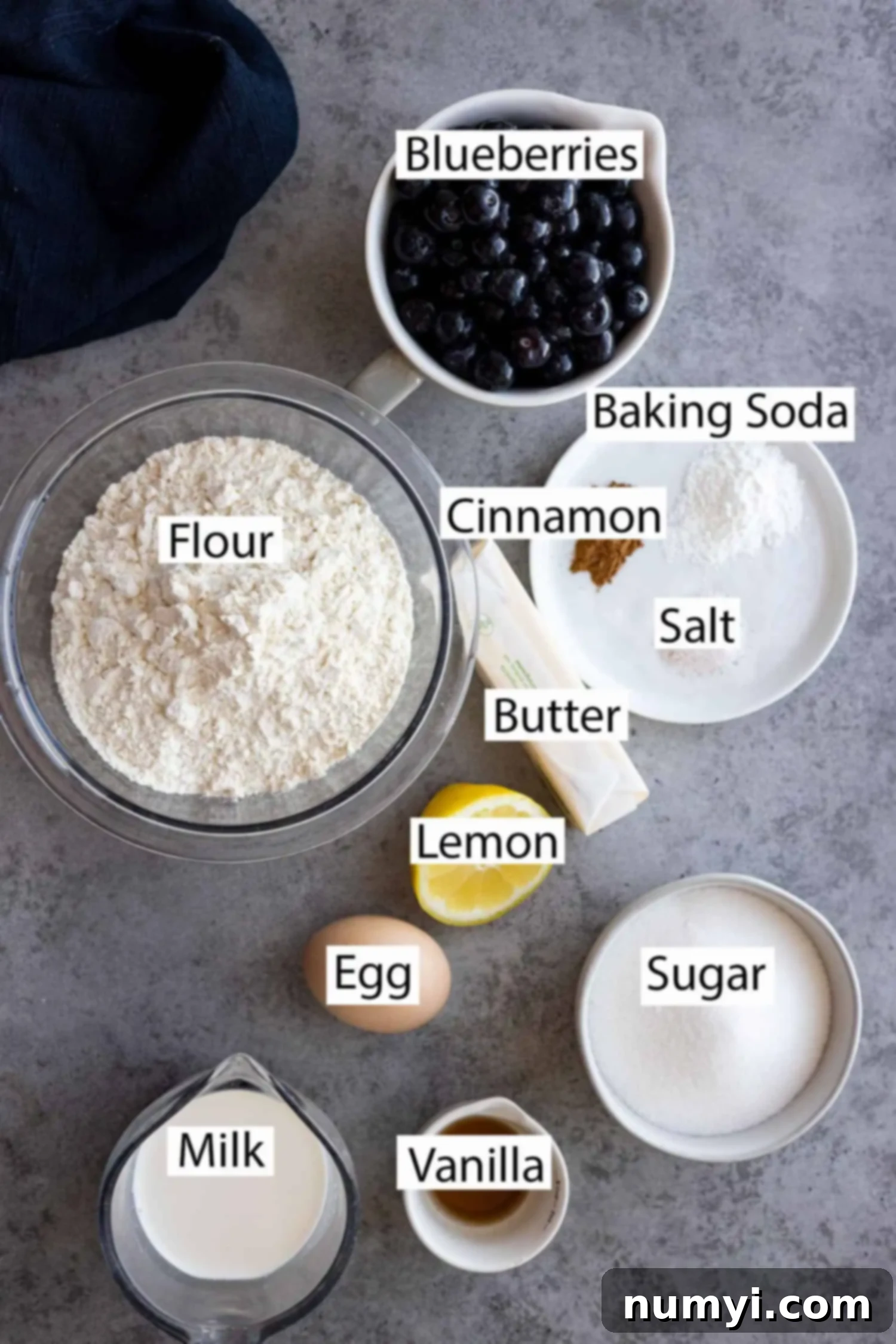 A flat lay of all the essential ingredients for blueberry buckle, including fresh blueberries, flour, sugar, butter, eggs, milk, vanilla, baking powder, cinnamon, and salt.