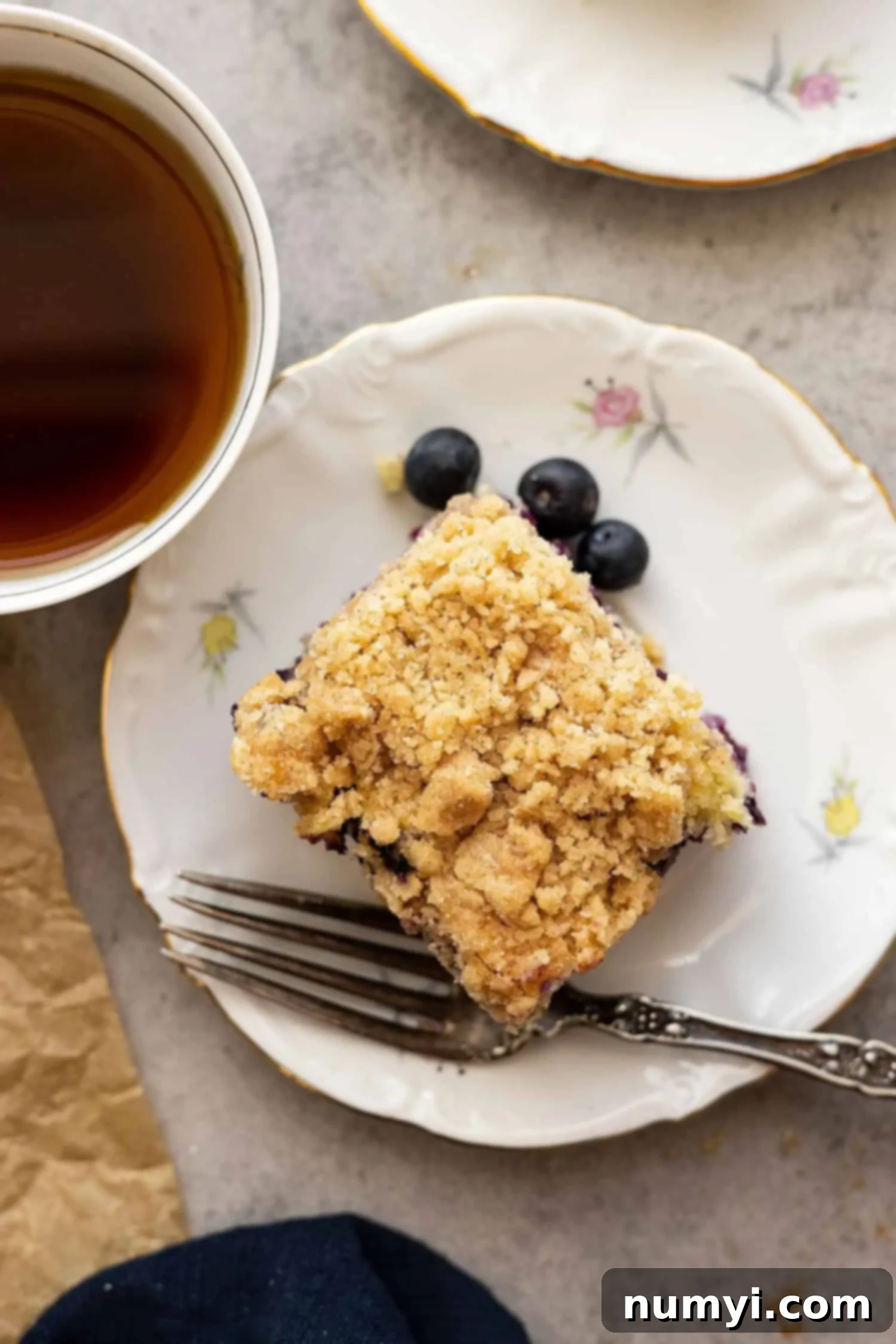 An overhead shot of a slice of blueberry buckle on a plate, with a fork and a cup of coffee off to the side, ready for a cozy breakfast.
