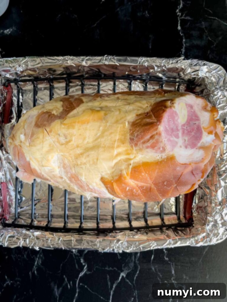A scored ham, flat side down, resting on a roasting rack inside a foil-lined roasting pan, ready for baking.