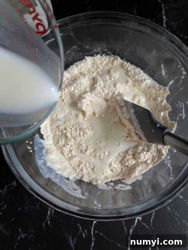 Effortless Butter Swim Biscuits 5 Buttermilk being gently stirred into the dry ingredients in a clear glass mixing bowl, forming a shaggy dough.