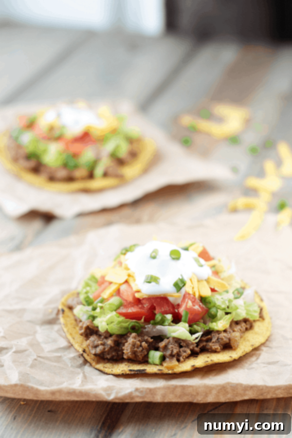 a tostada shell on a piece of parchment paper on a wood surface with beef and bean mixture on top of it, along with lettuce, tomato, cheese, and sour cream