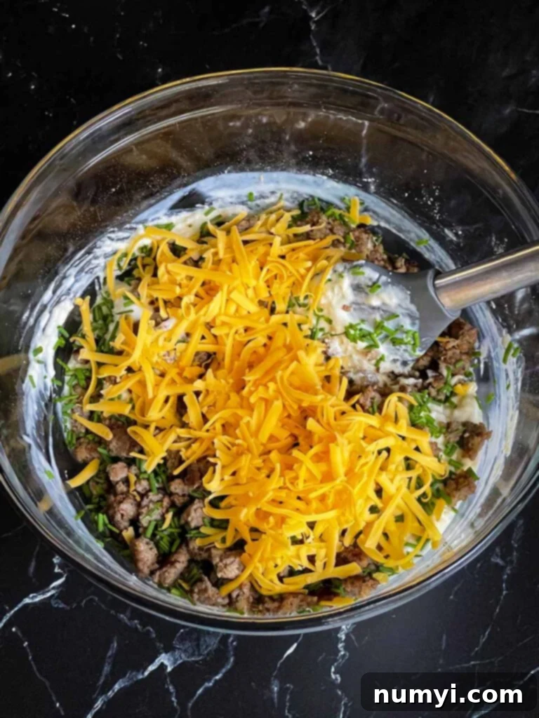 Cooked sausage, shredded cheese, and chives being stirred into the batter with a silver spatula.