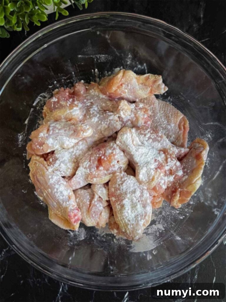 Raw chicken wings in a clear glass mixing bowl, generously sprinkled with white baking powder, ready for coating to enhance crispiness.