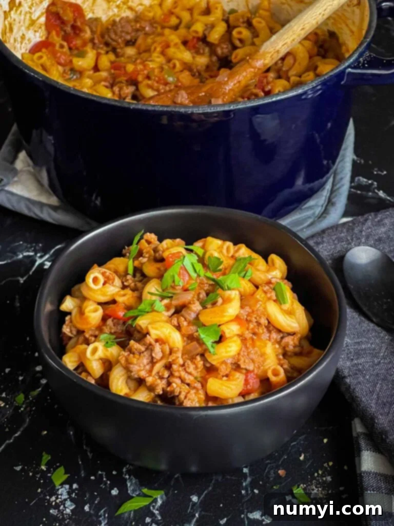 Grandma's American Goulash 3 A bowl of american goulash in a dark bowl with a dutch oven full of goulash in the background.
