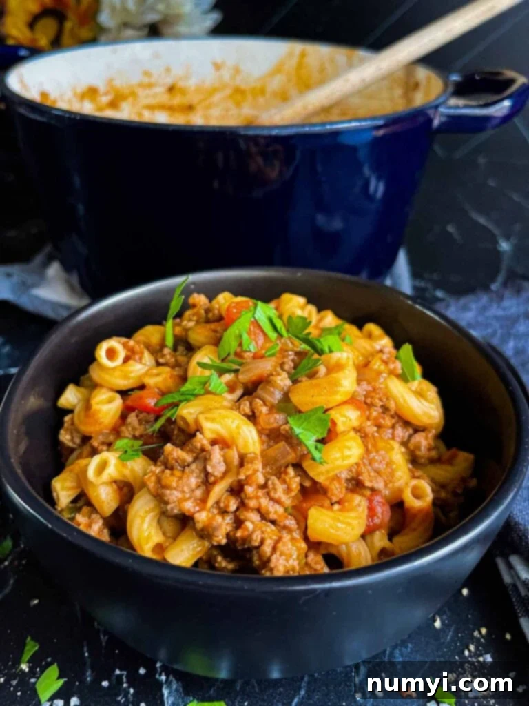Grandma's American Goulash 8 A bowl of American goulash (old fashioned goulash) in a dark bowl with a dutch oven in the background.