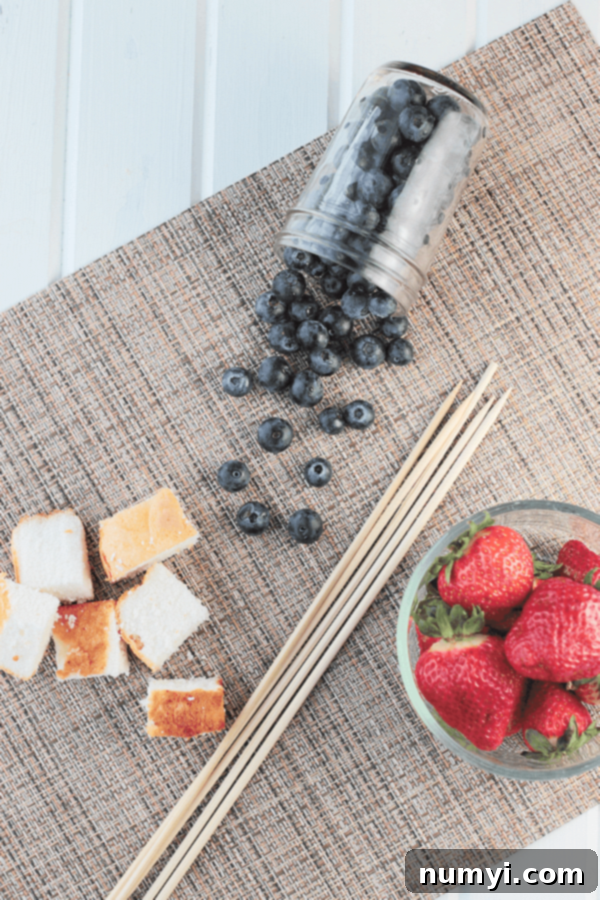 a bowl of strawberries, a jar of blueberries, wooden skewers, and chunks of angel food cake on a brown woven surface