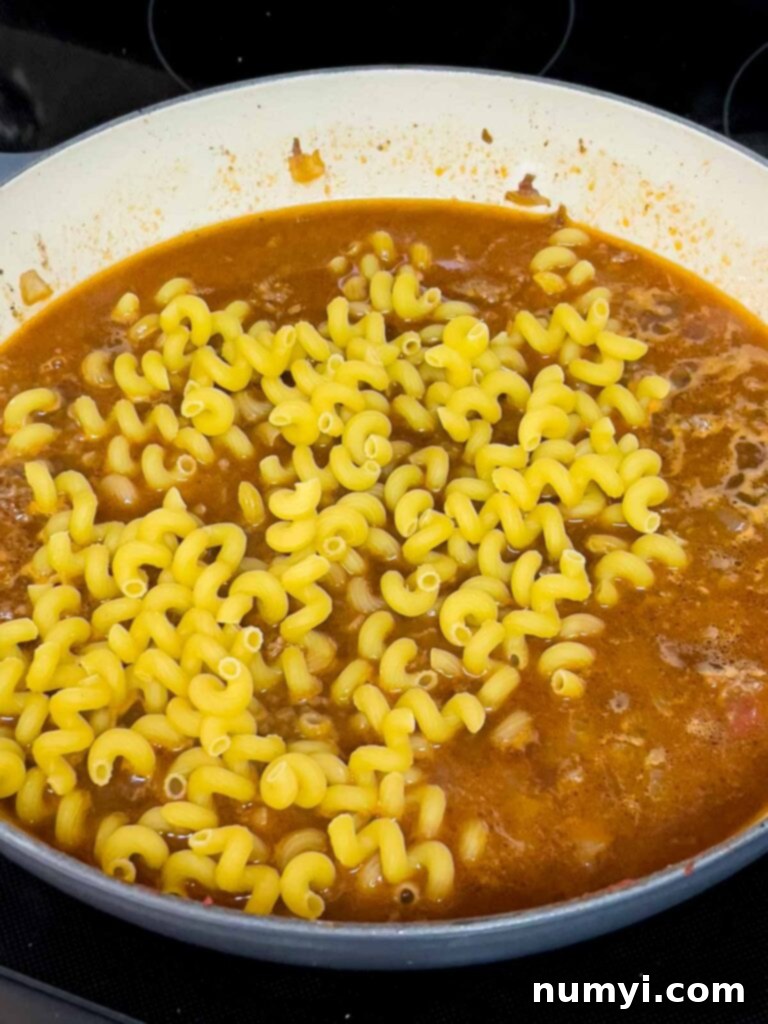 Uncooked Cavatappi pasta added to the beef and stock mixture in a cast iron skillet, before simmering.