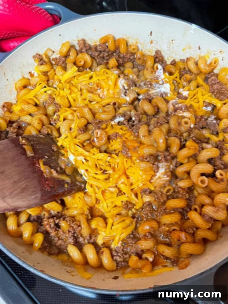 The creamy, cheesy sauce being mixed into the cooked ground beef and pasta in a cast iron skillet.