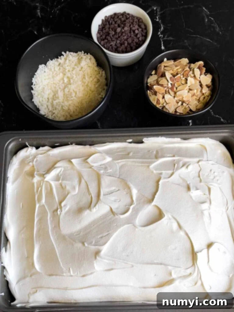 Whipped topping spread over the Almond Joy cake with coconut, almonds, and chocolate chips in the background.
