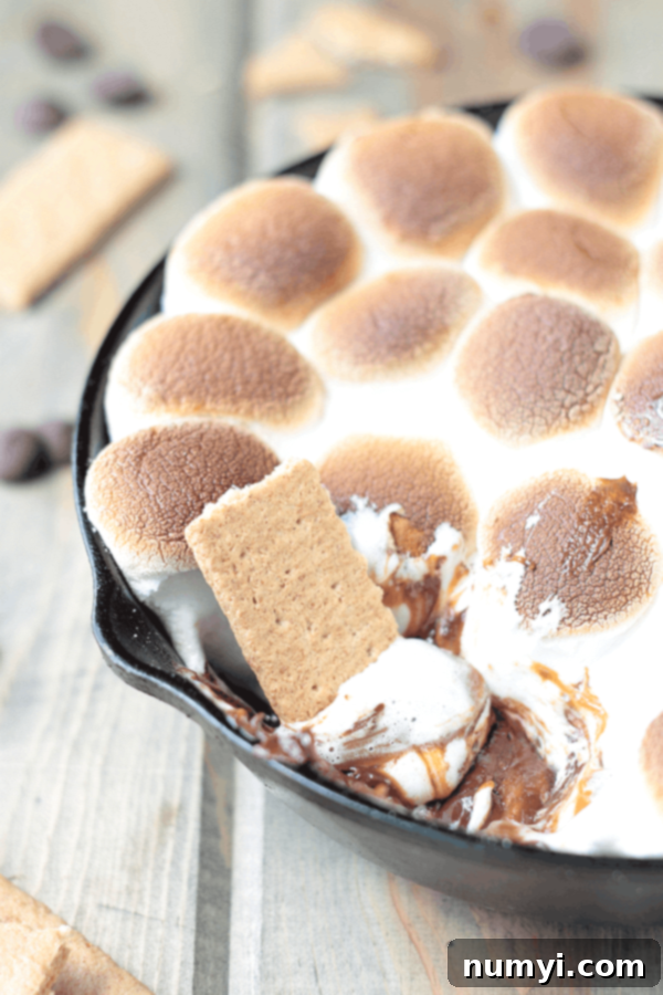 A black cast iron skillet brimming with toasted marshmallows atop a melted caramel and chocolate s'mores dip, with a graham cracker dipping in