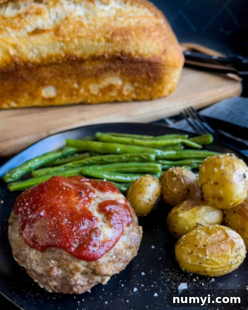 Mini meatloaf, roasted potatoes and green beans on a dark plate with a loaf of sourdough bread behind it.
