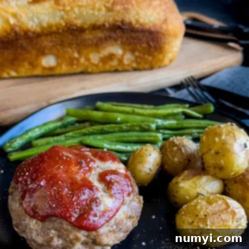Mini meatloaf, roasted potatoes and green beans on a dark plate with a loaf of sourdough bread behind it.