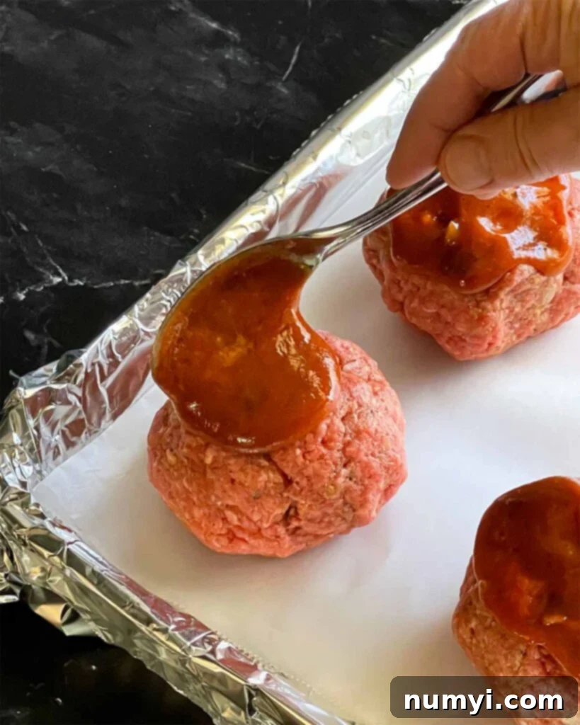 Unbaked mini meatloaves on a foil lined baking sheet with the sauce being spooned on top of them.