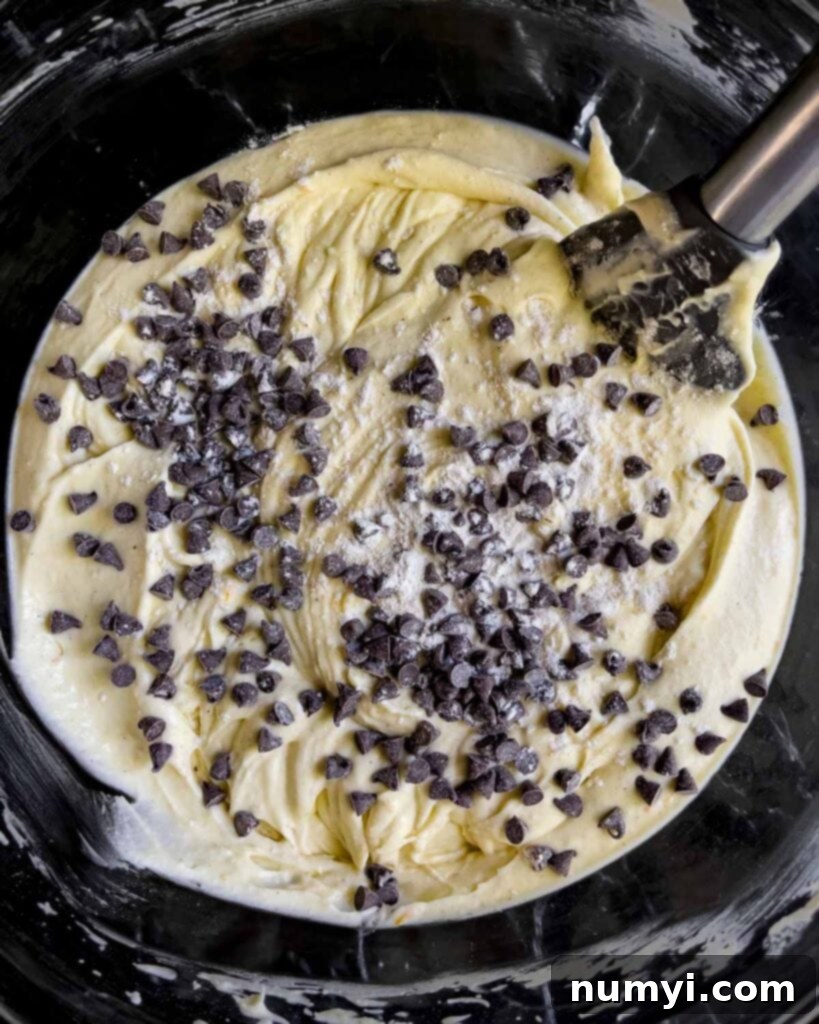 Flour-dusted mini chocolate chips being gently folded into the smooth cannoli cheesecake batter with a spatula.