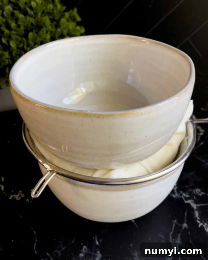 Ricotta cheese being drained in cheesecloth within a strainer, placed over a bowl with another bowl weighing it down for maximum moisture removal.