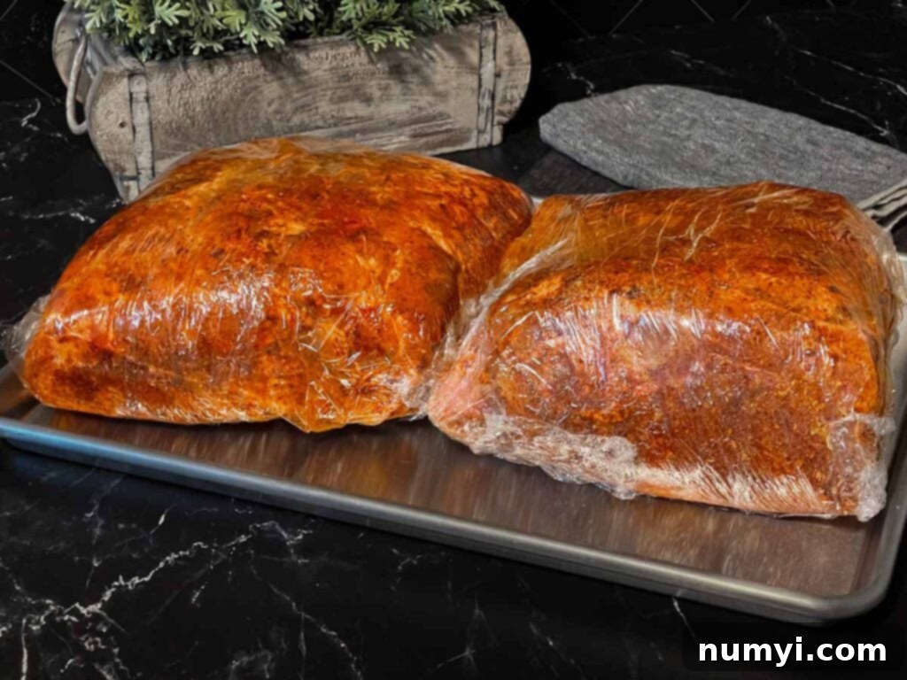 Two pork butts with rub applied, wrapped in plastic wrap.