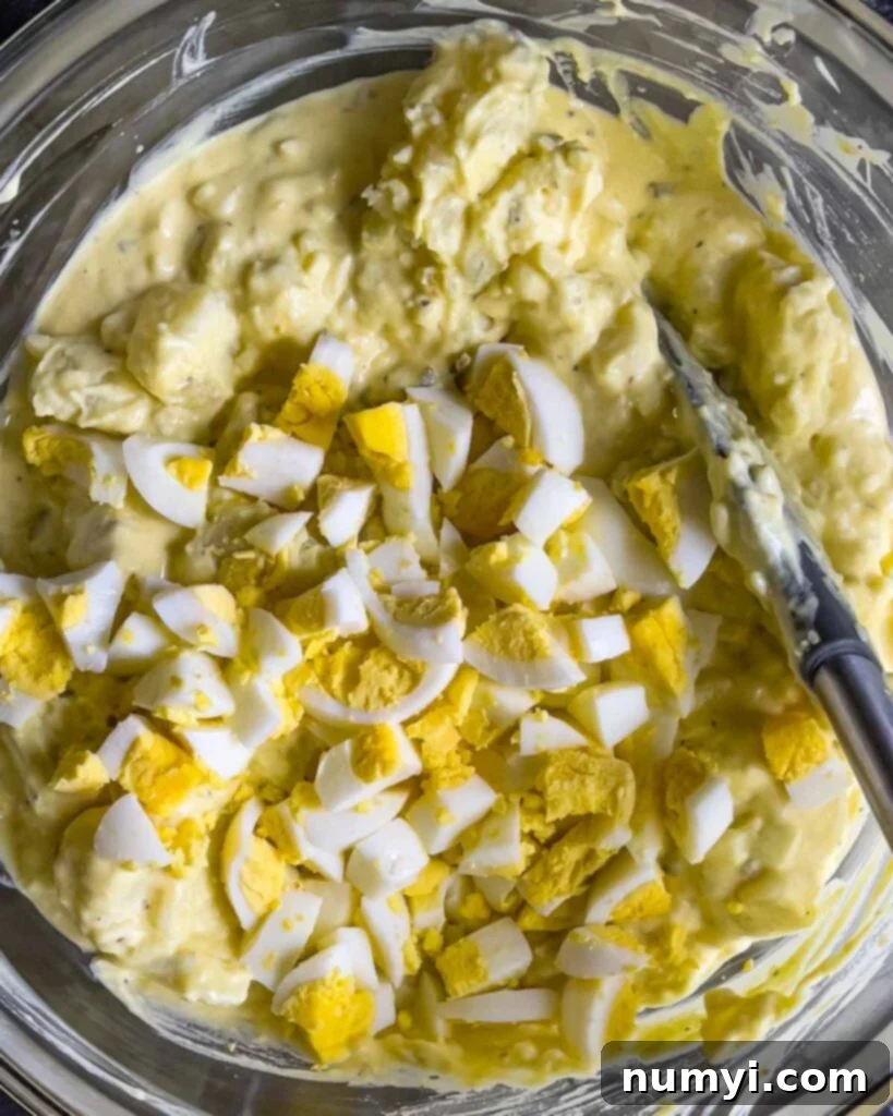 Chopped hard-boiled eggs being added to the Southern potato salad mixture in a glass mixing bowl.