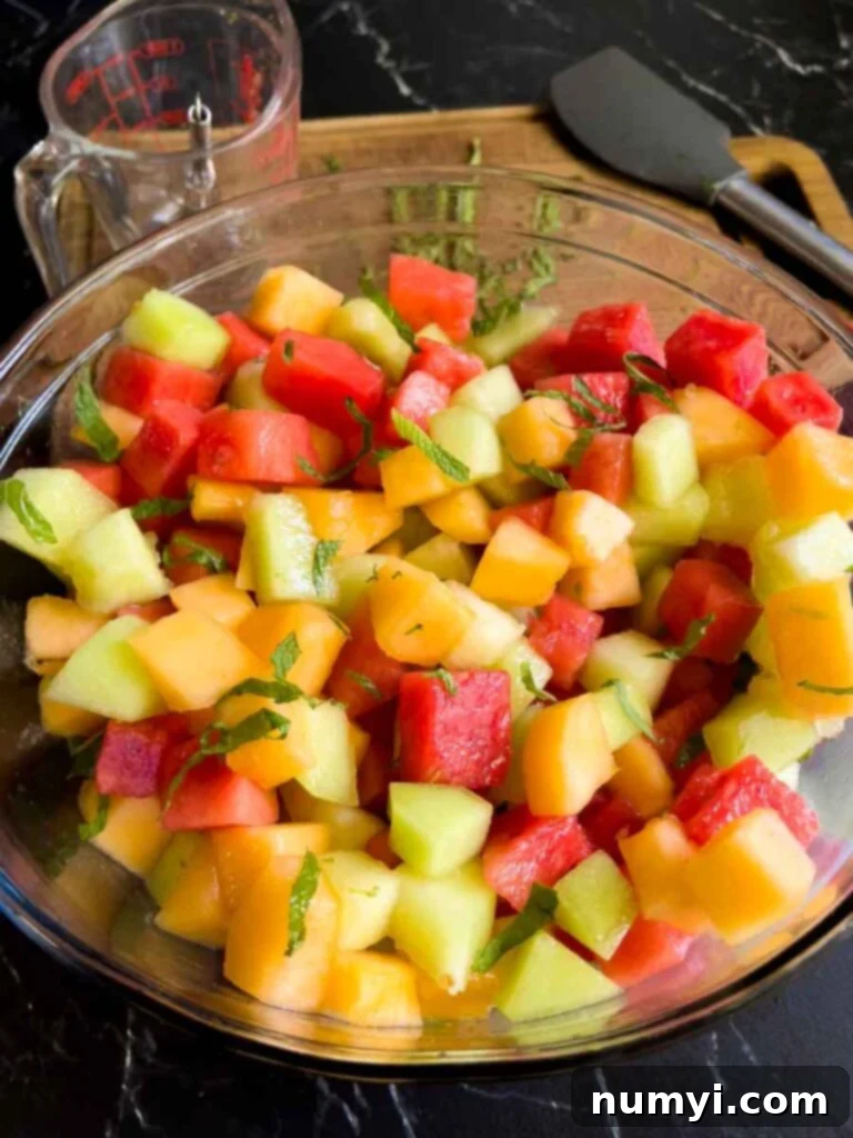 Mixed Melon Fruit Salad with Honey Lime and Mint 9 The freshly dressed mixed melon fruit salad, glistening with its honey-lime mint coating, presented in a large glass mixing bowl on a dark surface, ready to be served.