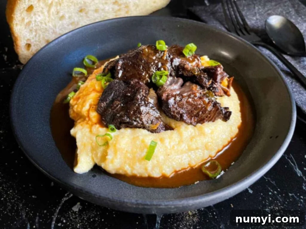 Braised beef cheek on a bed of polenta, garnished with fresh green onion. The dish is presented in a white bowl with a spoon.