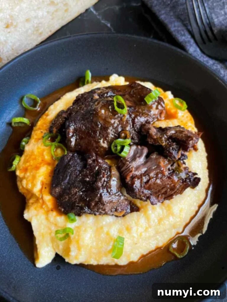 A braised beef cheek resting on a bed of creamy polenta, garnished with thinly sliced green onion, highlighting the incredibly tender texture of the meat.