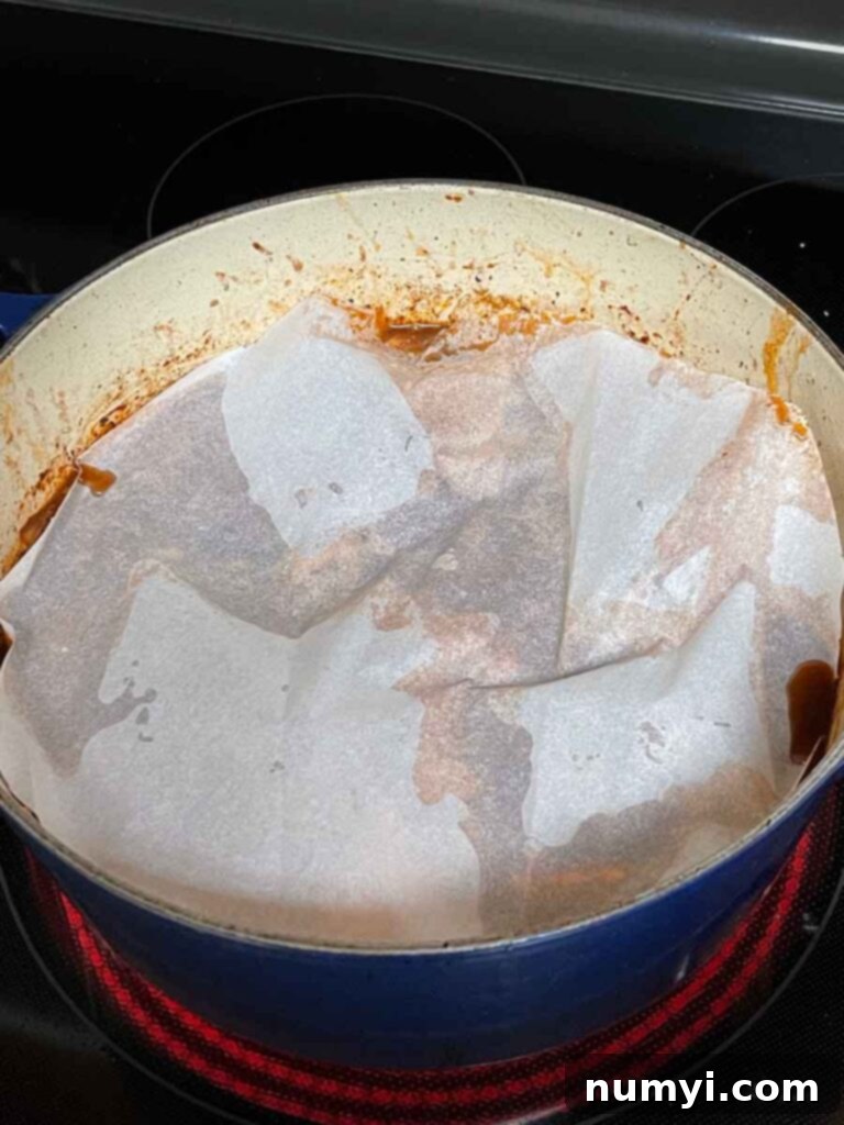 Braised beef cheeks simmering in a Dutch oven, covered with a round piece of parchment paper (cartouche) to ensure even cooking.
