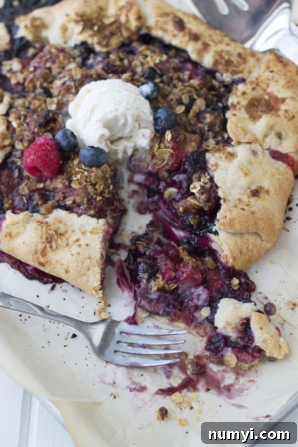 A close-up shot of a slice of berry galette on a white plate, revealing the vibrant, juicy berry filling and the golden, flaky crust, with a fork resting beside it and a dollop of melting vanilla ice cream on top.