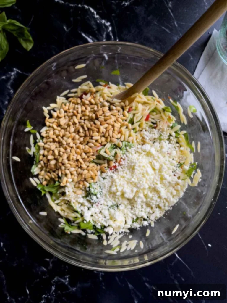 Toasted pine nuts and crumbled feta cheese added to the spinach and feta orzo salad in a glass bowl on a dark surface.