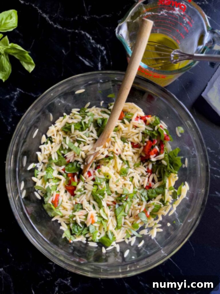 Vegetables added to the orzo in a glass bowl with orzo salad dressing in a glass measuring cup.