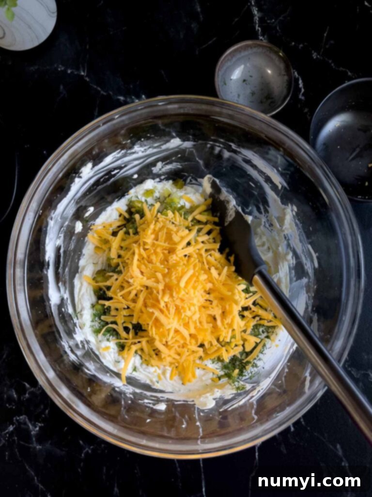Cheddar cheese, dill, and diced dill pickles added to the cream cheese mixture in a glass bowl on a dark surface.
