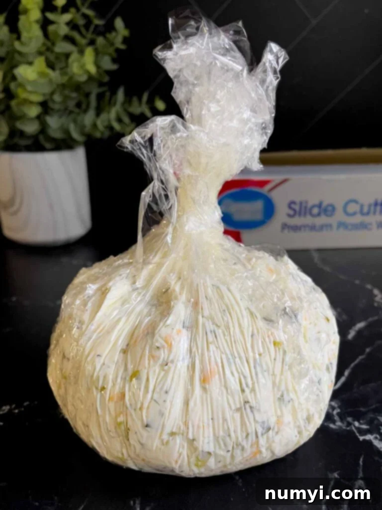 Dill pickle cheese ball wrapped in plastic wrap on a dark surface.