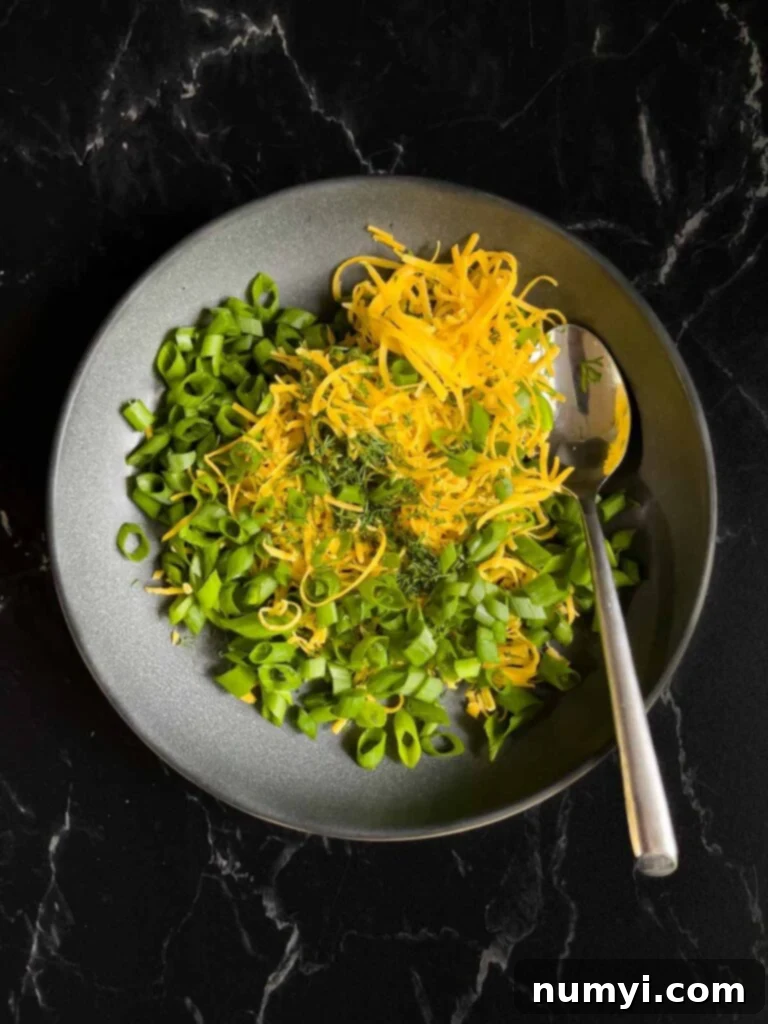 Green onions, dill, and cheddar cheese toppings for the dill pickle cheese ball in a dark bowl on a dark surface.