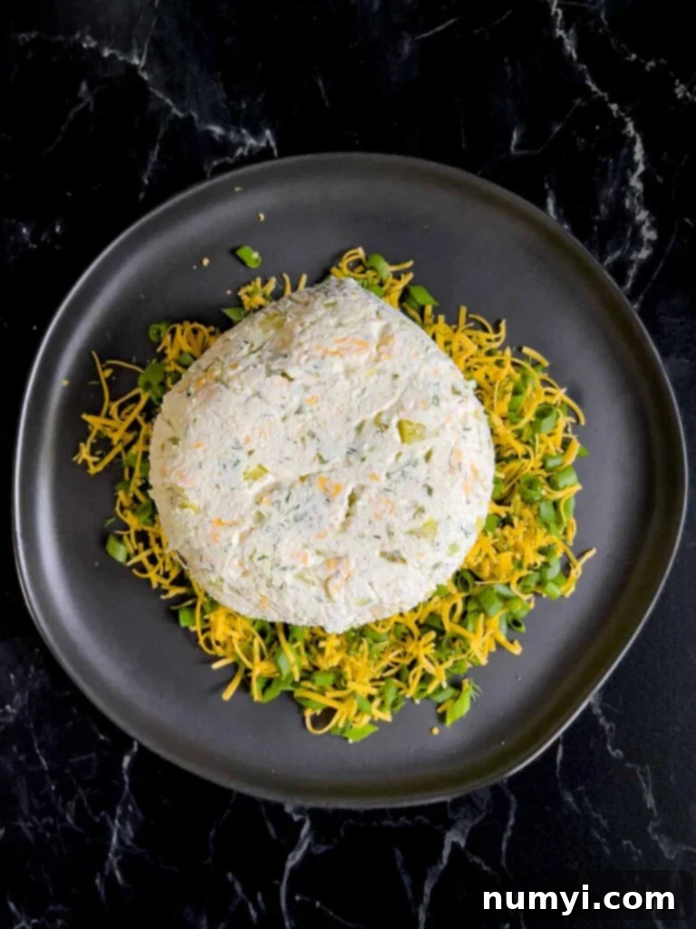 Dill pickle cheese ball on a bed of garnish of cheddar cheese, dill, and green onions on a dark plate on a dark surface.
