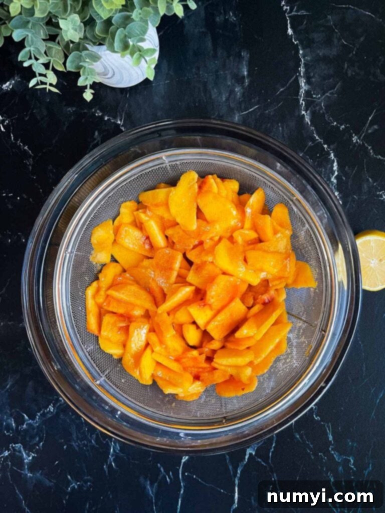 Sliced and chunked peaches releasing their juices in a fine mesh strainer over a large glass bowl.